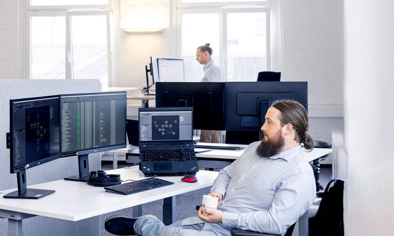 Foto vom TLK-Thermo Büro, ein Kollege sitzt entspannt bei einer Kaffeepause am Schreibtisch. Im Hintergrund sieht man Computerbildschirme, auf denen die MoBA-Automation läuft.